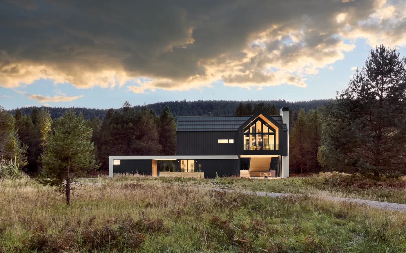 M&oslash;rk enebolig i skoglandskap under dramatisk himmel med varm gavl og store glassfelt.