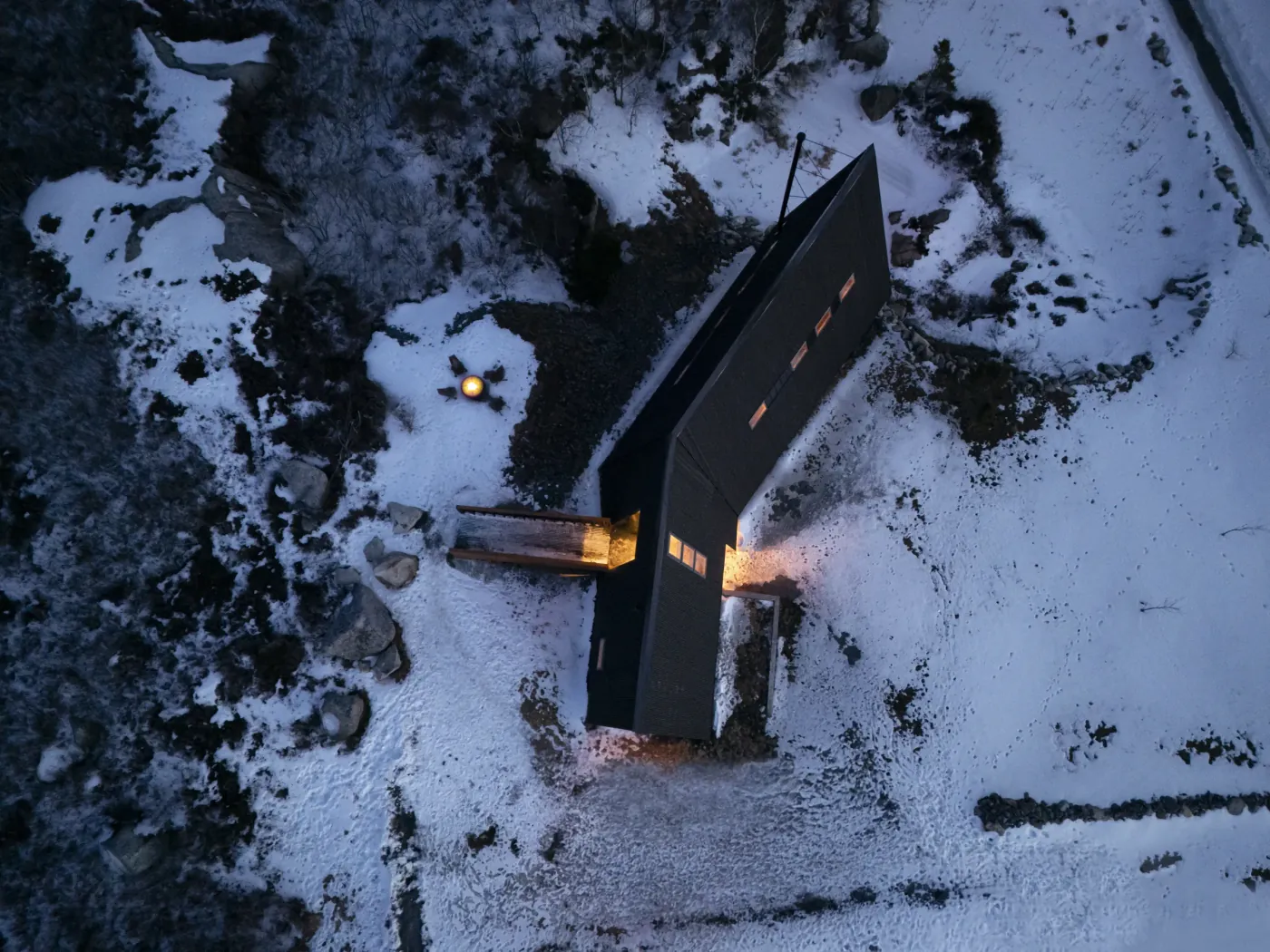 Fugleperspektiv av A-hytte i vinterterreng med opplyst inngang og smal grunnflate.